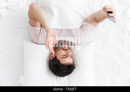 Ansicht von oben mit einem lächelnden gutaussehenden jungen Mann im Bett, mit Handy Stockfoto
