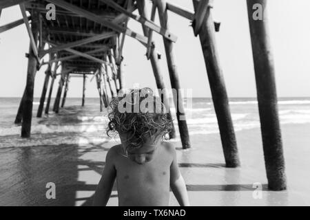 Schwarz-weiß Foto eines Toddler Boy mit lockigem Haar, als er unter einem Pier am Meer im Surfside Beach, SC steht. Stockfoto