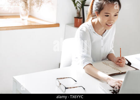 Lächelnd Asian Business Frau etwas Schreiben und Verwenden von Laptop während der Sitzung durch die Tabelle im Büro Stockfoto