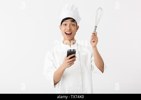 Asiatische Koch tragen Uniform, die auf weißem Hintergrund aufgeregt, über Handy, mit Schneebesen Stockfoto