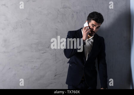 Eine ernsthafte hübscher junger Geschäftsmann in einem schwarzen Anzug über sein Telefon vor einer grauen Wand in einem Studio. Stockfoto