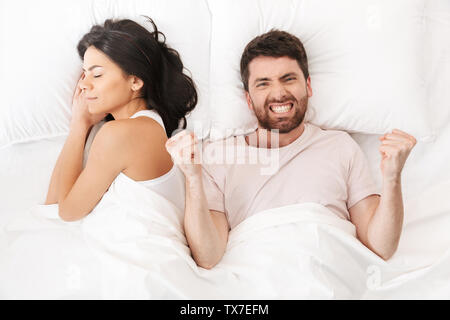 Bild eines glücklichen neugierige junge Mann liegt im Bett unter der Decke in der Nähe der schlafenden Frau Sieger Geste machen. Stockfoto