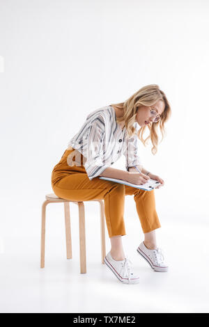 Foto des europäischen Denkens Frau Brille im Stuhl sitzen und schreiben auf Zwischenablage über weissen Hintergrund im Studio isoliert Stockfoto