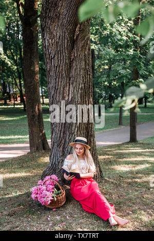 Frau in vintage Kleid und Strohhut auf dem Gras lesen Buch im Sommer Wald sitzen Stockfoto