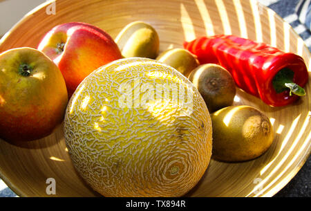 Close up Bambus Obstkorb mit Melone, Äpfel, Kiwis, Paprika von Abendsonne Balken leuchtet auf Stockfoto