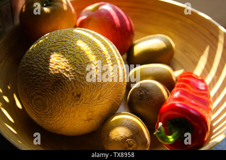 Close up Bambus Obstkorb mit Melone, Äpfel, Kiwis, Paprika von Abendsonne Balken leuchtet auf Stockfoto