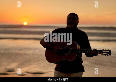 Gerne älterer Mann spielt eine akustische Gitarre am Strand bei Sonnenuntergang. Stockfoto