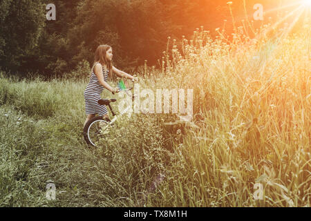 Mädchen in gestreiften Kleid Reiten Fahrrad. Die Zeit im Freien. Kinder spielen draußen im Frühjahr, im Sommer. Kindheitserinnerungen. Wandern im Park, Wald. Das Gehen auf Reise, Abenteuer in der Wildnis Stockfoto