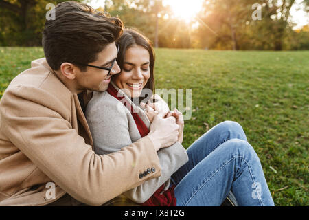 Bild von brunette kaukasischen Paar lächelnd und umarmen einander beim Sitzen auf Gras im Park Stockfoto