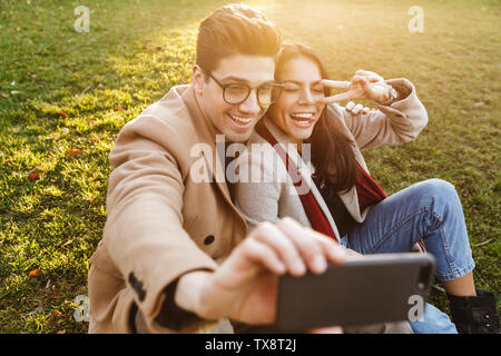 Image der jungen europäischen Paar Mann und Frau 20 s unter selfie auf Handy und zeigt Frieden singen während der Sitzung auf Gras im Park Stockfoto