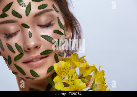 In der Nähe von Frau Gesicht voller grüne Blätter mit gelben Blumen, die isoliert auf grau, Mädchen mit ihren Augen von florals geschlossen umgeben Stockfoto