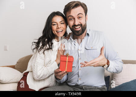 Bild des charmanten Paar Mann und Frau mit Geburtstagsgeschenk box beim Sitzen auf einem Sofa im hellen Zimmer zu Hause Stockfoto