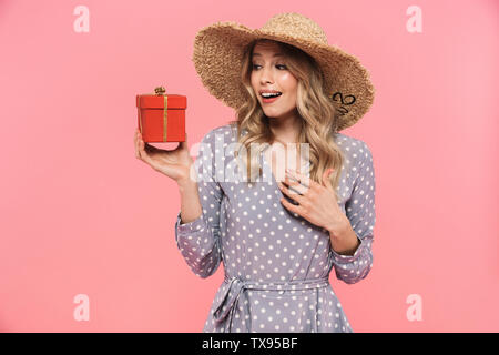 Schöne junge blonde Frau Sommer Hut und Kleid stehen über rosa Hintergrund isoliert, kleines Geschenk Box Stockfoto