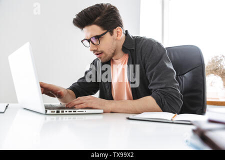 Seitenansicht des Schockiert ernste stattlicher Mann in Brillen mit Laptop Computer, während durch die Tabelle im Büro sitzen Stockfoto