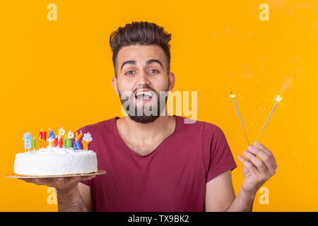 Positive junge Mann hält ein Happy birthday cake und zwei brennende bengalische Lichter auf gelbem Hintergrund posiert. Stockfoto