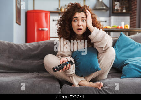 Foto von betonte junge Frau mit Haus Kleidung sitzt auf einem Sofa in der Wohnung und Fernbedienung Stockfoto