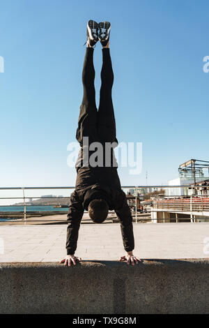 Foto von Parkour Kerl 20 s in schwarz Trainingsanzug, Akrobatik und stehend auf seine Arme während der morgendlichen Workout von Seaside Stockfoto