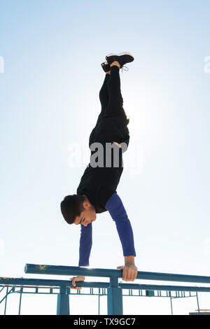 Foto sportlicher Kerl 20 s in schwarz Trainingsanzug, Akrobatik auf horizontale Gymnastik bar während der morgendlichen Workout von Seaside Stockfoto