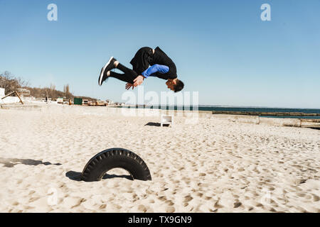 Foto von bold Kerl 20 s in schwarz Trainingsanzug, Parkour und springen während der Morgen Akrobatik von Seaside Stockfoto