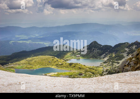 Gerahmte Blick auf die sonnenbeschienenen Fish Lake und das Kleeblatt See, zwei der berühmten sieben Rila Seen, durch Vordergrund Schnee und Berge Stockfoto