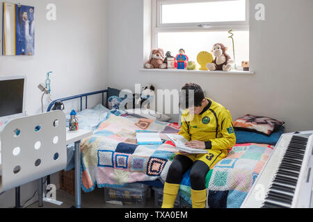 England, Uk-Young Junge, 11 Jahre in der kleinen Liga Fußball-Outfit liest ein Buch in seinem Schlafzimmer Stockfoto