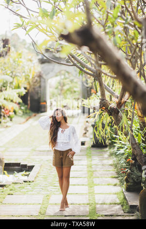 Schlanke brünette Frau in weißem Kittel und braune Hosen im marokkanischen Stil wandern im tropischen Garten Stockfoto