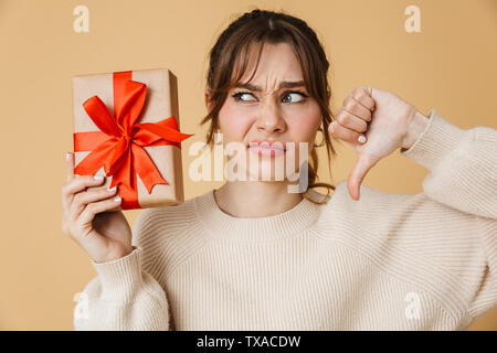 Schöne umgekippt junge Frau trug Pullover stehend über beigen Hintergrund isoliert, Geschenkbox, Daumen nach unten Stockfoto