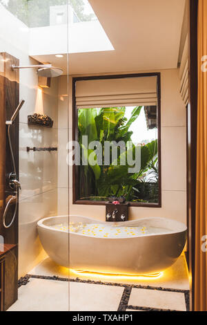 Moderne Einrichtung der Badezimmer mit Badewanne und tropischen Garten Blick durch das Fenster. Blütenbad mit Schaum blasen und gelbe Blumen in Spa Stockfoto