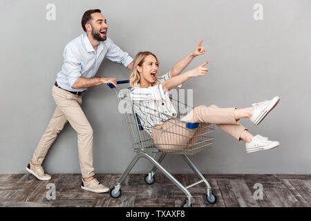 Image der Jungen glückliche Menschen in Freizeitkleidung drücken Warenkorb mit blonden Frau über graue Wand isoliert Stockfoto