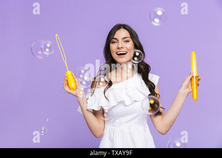 Schöne fröhliche junge Frau Sommer Outfit stehen über violette Hintergrund isoliert, bläst Seifenblasen Stockfoto