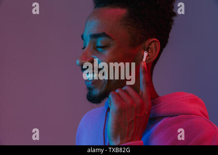 Portrait von entspannt afrikanische amerikanische Mann in bunten hoodie Musik hören mit earpod über violett Hintergrund isoliert Stockfoto
