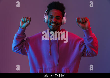 Portrait von Angeregten afrikanische amerikanische Mann in bunten hoodie Musikhören mit Kopfhörern über violett Hintergrund isoliert Stockfoto