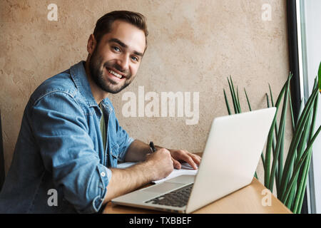 Foto junger bärtiger Mann tragen Jeanshemd schreiben mit Laptop im Cafe drinnen arbeiten Stockfoto