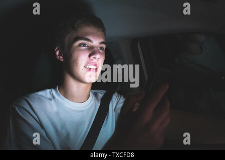 Abgelenkt Teenager, der ein Auto fährt mit seinem Handy in der Hand. Das Licht vom Bildschirm des Telefons ist erhellend sein Gesicht. Stockfoto