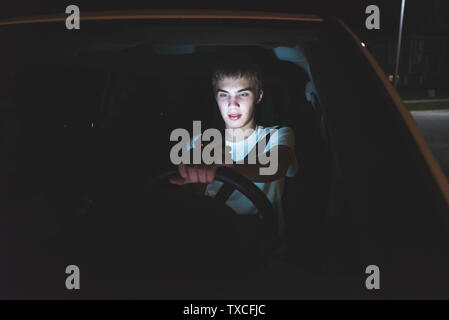 Abgelenkt Teenager, der ein Auto fährt mit seinem Handy in der Hand. Das Licht vom Bildschirm des Telefons ist erhellend sein Gesicht. Stockfoto