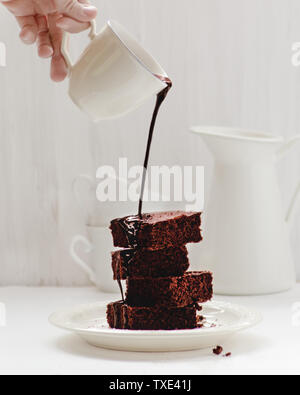 Chocolate Brownie mit Schokoladensauce. Cluse. Weißer Hintergrund, weiße Requisiten, weiße Tassen, weiße Platte. Sirup Schokolade. Hand gießt. Stockfoto