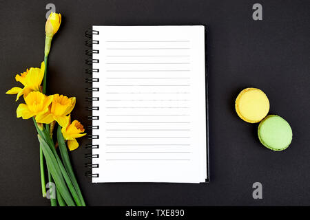 Ansicht von oben flach leer Notebook legen mit Frühlingsblumen Narzissen und Makronen Dessert auf schwarzem Hintergrund. Text Raum. Nach doi Liste. Stockfoto