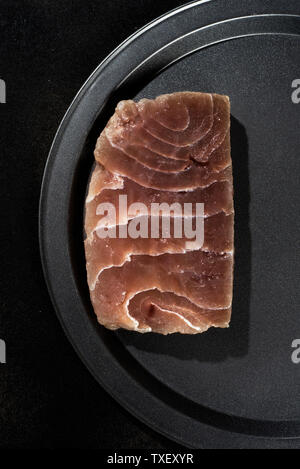 Hohe Betrachtungswinkel eines rohen Thunfisch Steak auf einem schwarzen Fach, auf einer schwarzen Fläche platziert Stockfoto