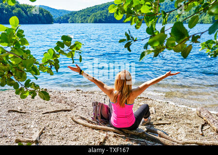 Glückliche junge Frau mit ihren Armen in der Freude verbreiten, sitzen auf einem Berg Lake Shore, mit ihrem Rucksack in der Nähe von ihr und trägt einen hellen fuchsia Shirt. Sunn Stockfoto