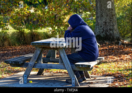 Eine Person mit einer Haube sitzt an einem Tisch für Picknick im Park Stockfoto