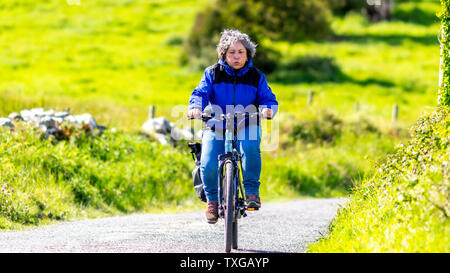 Müde reife mexikanische Frau, die ihr Fahrrad auf einem Hügel, Ausdruck auf ihrem Gesicht, ausatmen, Luft, wunderbar sonniger Frühling im Dorf Doolin in Stockfoto