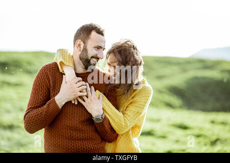 Portrait von ein paar schöne in hellen Pullover umarmen auf der grünen Wiese, während in den Bergen reisen gekleidet Stockfoto