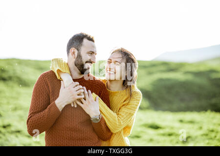 Portrait von ein paar schöne in hellen Pullover umarmen auf der grünen Wiese, während in den Bergen reisen gekleidet Stockfoto