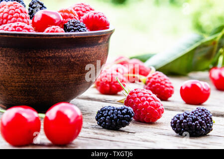 Saftige frische leckere Beeren, Kirschen, Himbeeren, Maulbeeren in einer Schüssel auf Holzbretter auf einem Hintergrund von grünem Gras. Close-up Stockfoto