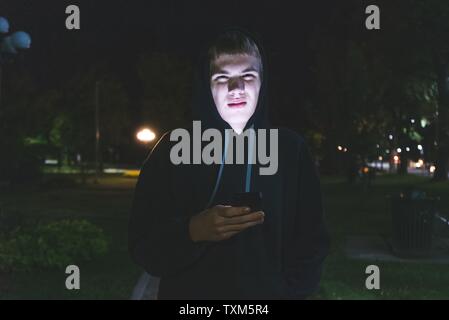 Traurige Teenager auf seinem Handy beim Stehen in einem Stadtpark bei Nacht. Stockfoto