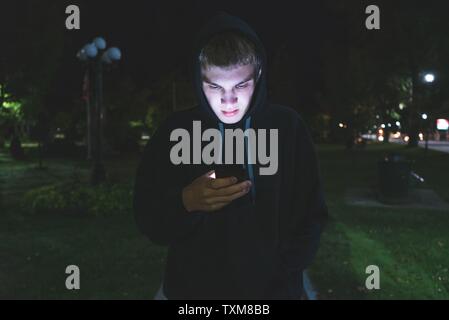 Traurige Teenager auf seinem Handy beim Stehen in einem Stadtpark bei Nacht. Stockfoto