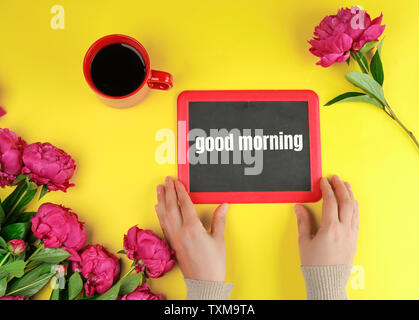 Zwei weibliche Hände halten eine schwarze Kreide Rahmen mit den Worten "Guten Morgen, rot Tasse mit schwarzem Kaffee und Strauß Pfingstrosen auf gelbem Hintergrund Stockfoto