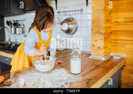 Eine kleine süße Mädchen fertig, einen Kuchen in der Küche zu backen. Rühren den Teig in eine Schüssel geben. Mehl und Milch auf den Tisch. Stockfoto