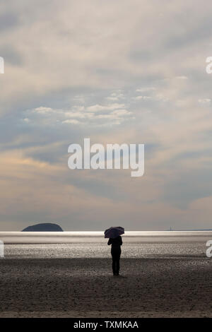 Entfernte einsame Figur mit Schirm isoliert auf leerem Strand Sand bei Sonnenuntergang, Weston-Super-Mare, Großbritannien. Stockfoto