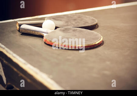 Zwei verschlissen roten und grauen Ping pong Schläger und einem weißen Kunststoff Kugel liegen auf einer alten grauen Holz- ping-pong Tisch Stockfoto